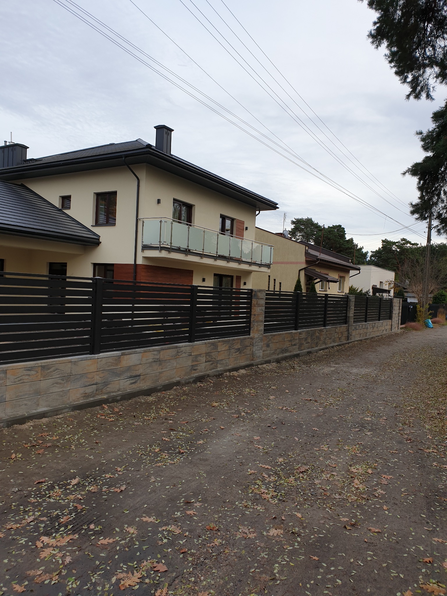 Nowoczesny, poziomy, czarny płot z podmurówką z jasnego kamienia na tle domu z balkonem i jesiennego krajobrazu.
