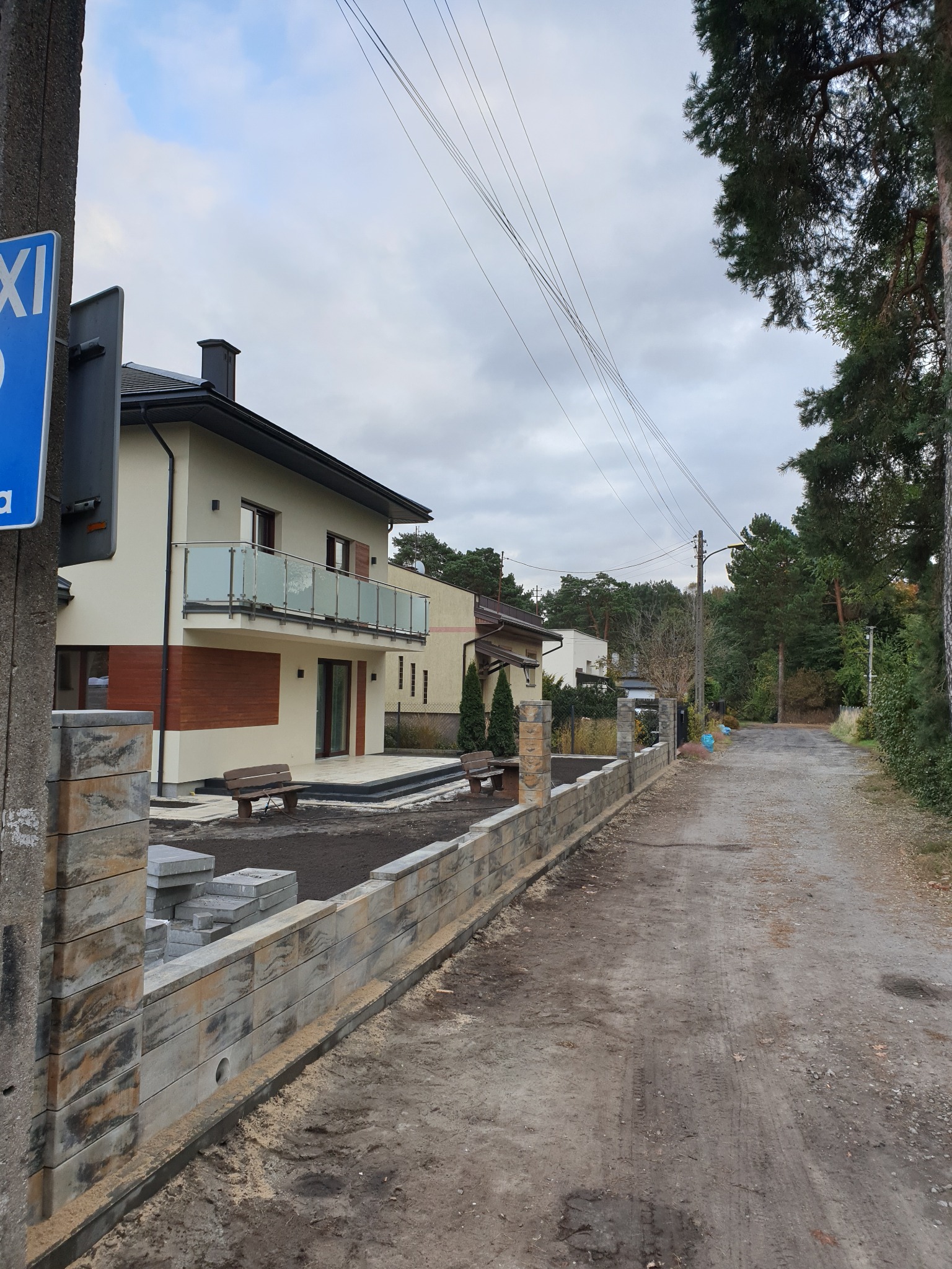 Wznoszenie ogrodzenia z betonowych bloczków o dekoracyjnej fakturze w sąsiedztwie nowoczesnego domu z balkonem, widok z polnej drogi.