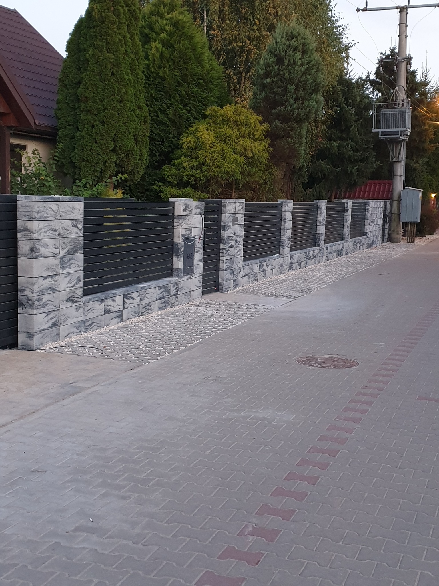 Nowoczesne ogrodzenie posesji z szarej kostki imitującej kamień i czarnych, poziomych paneli, z małą skrzynką gazową na jednym z filarów, widok z chodnika wyłożonego kostką.