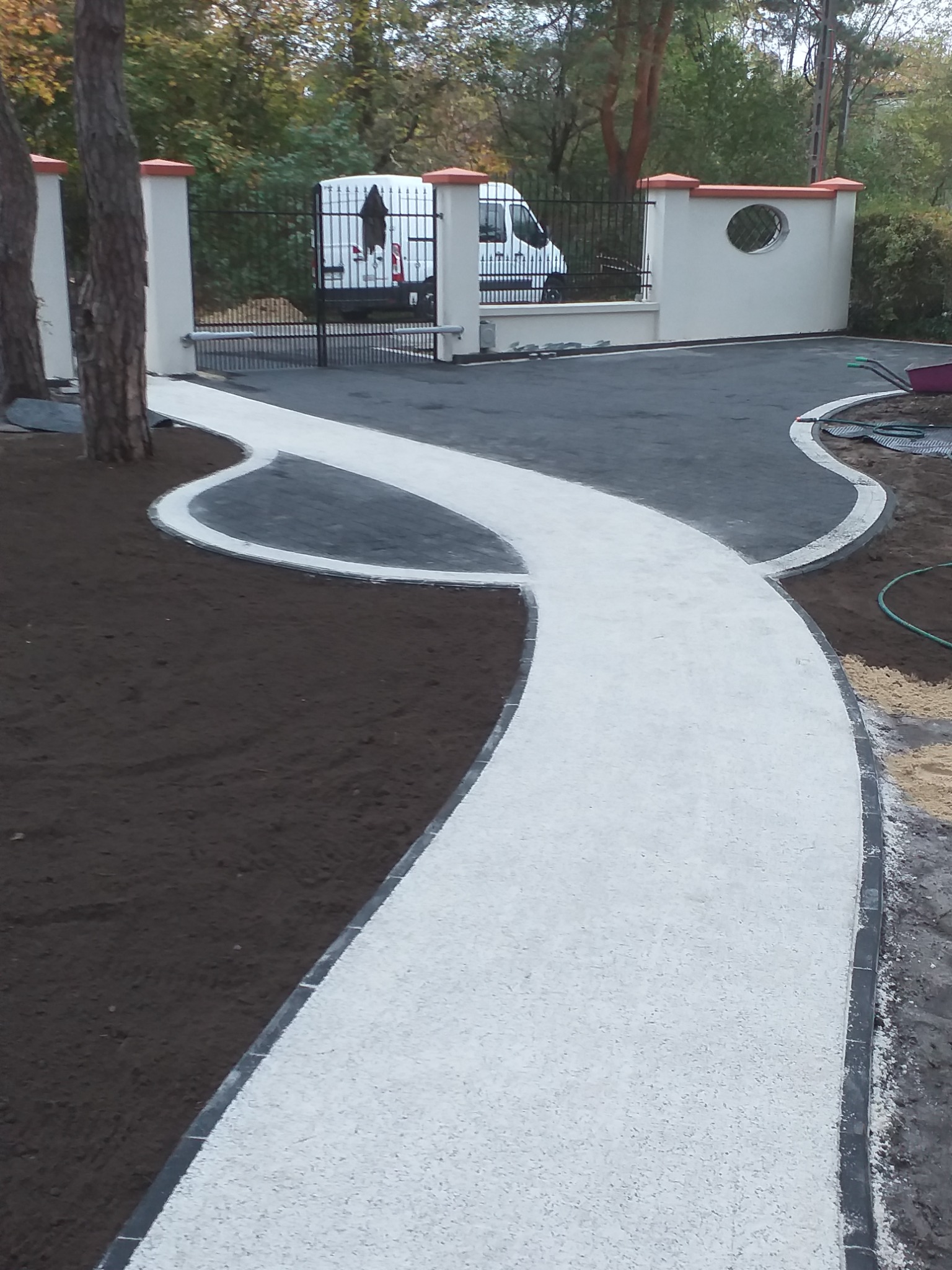 Ścieżka z jasnej kostki prowadząca do bramy wjazdowej z białymi słupkami, obok świeżo przygotowany teren pod trawnik.