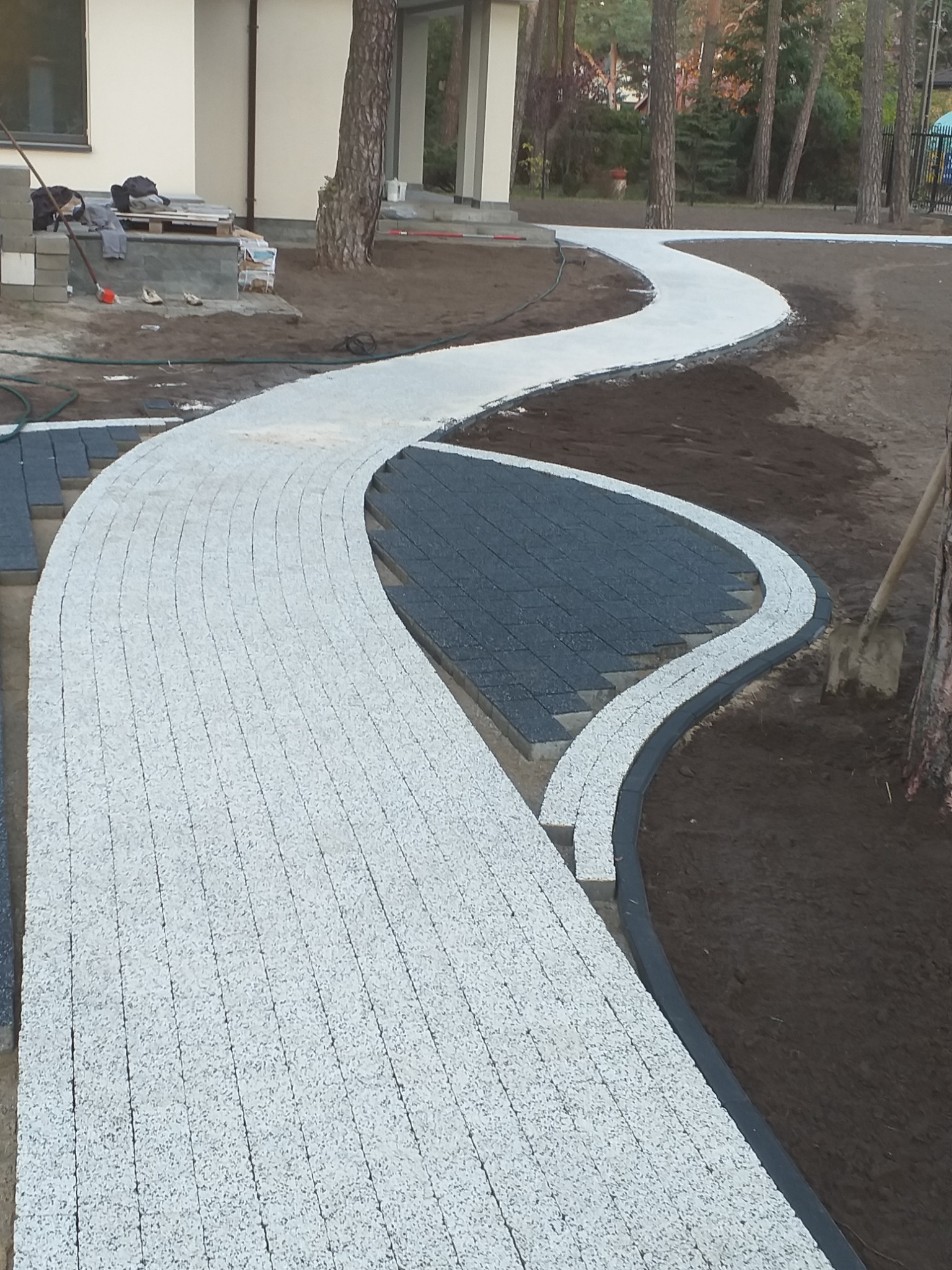 Kręta ścieżka z jasnej kostki brukowej w ogrodzie, obok widoczny fragment układanej ciemnej kostki i narzędzia pracy.