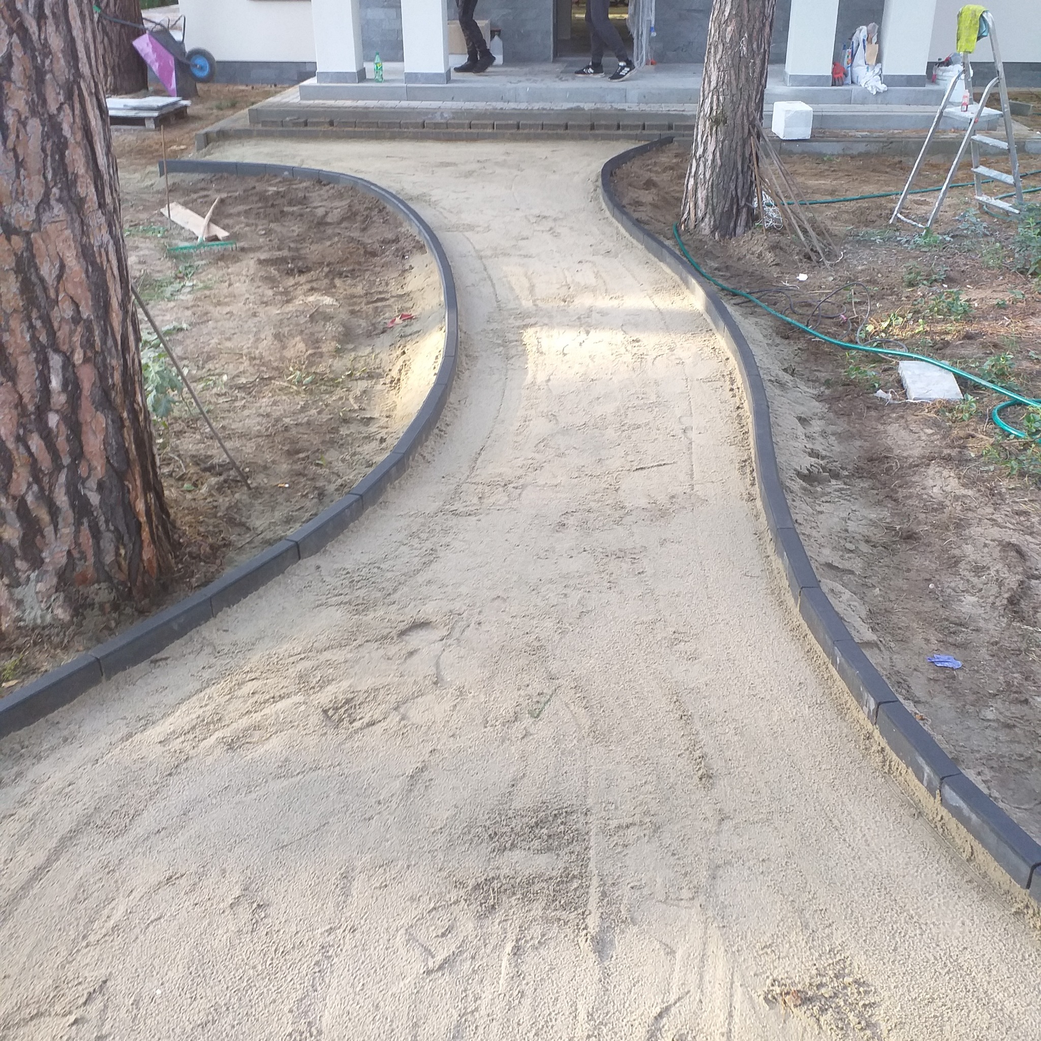 Przygotowanie podłoża pod ścieżkę z kostki, widoczne krawężniki z ciemnej kostki ułożone po łuku, piasek wyrównany, w tle budynek i drzewa.