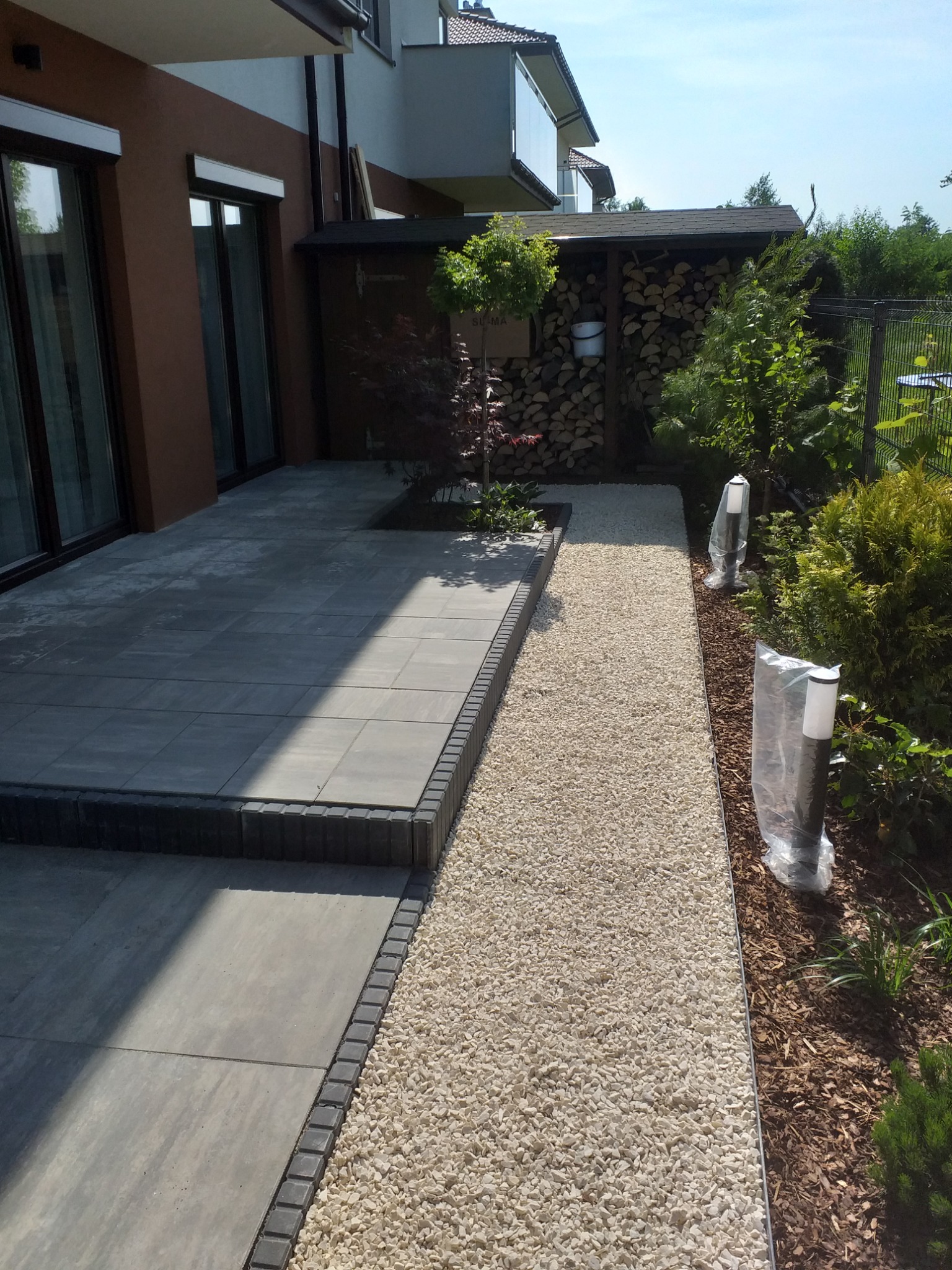 Nowoczesny taras z szarych płyt betonowych z obrzeżem z ciemnej kostki, przechodzący w ścieżkę z jasnego żwiru, oświetloną lampami ogrodowymi w foliowych osłonach, obok domu z brązową elewacją...