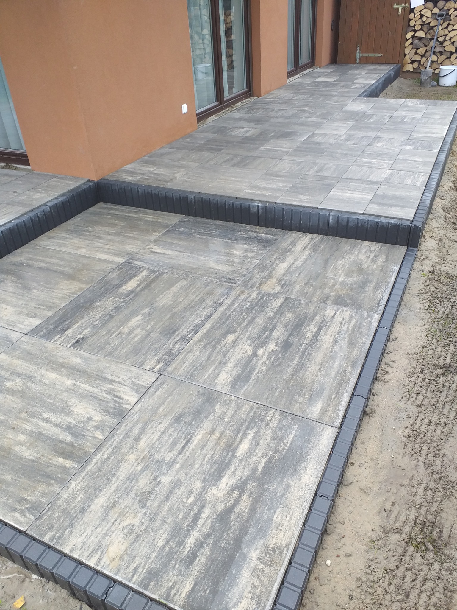 Nowoczesny taras z szarych, kwadratowych płyt z dekoracyjnym obrzeżem w kolorze grafitowym, prowadzący do drzwi balkonowych i drewnianego pomieszczenia gospodarczego z ułożonym drewnem opałowym.