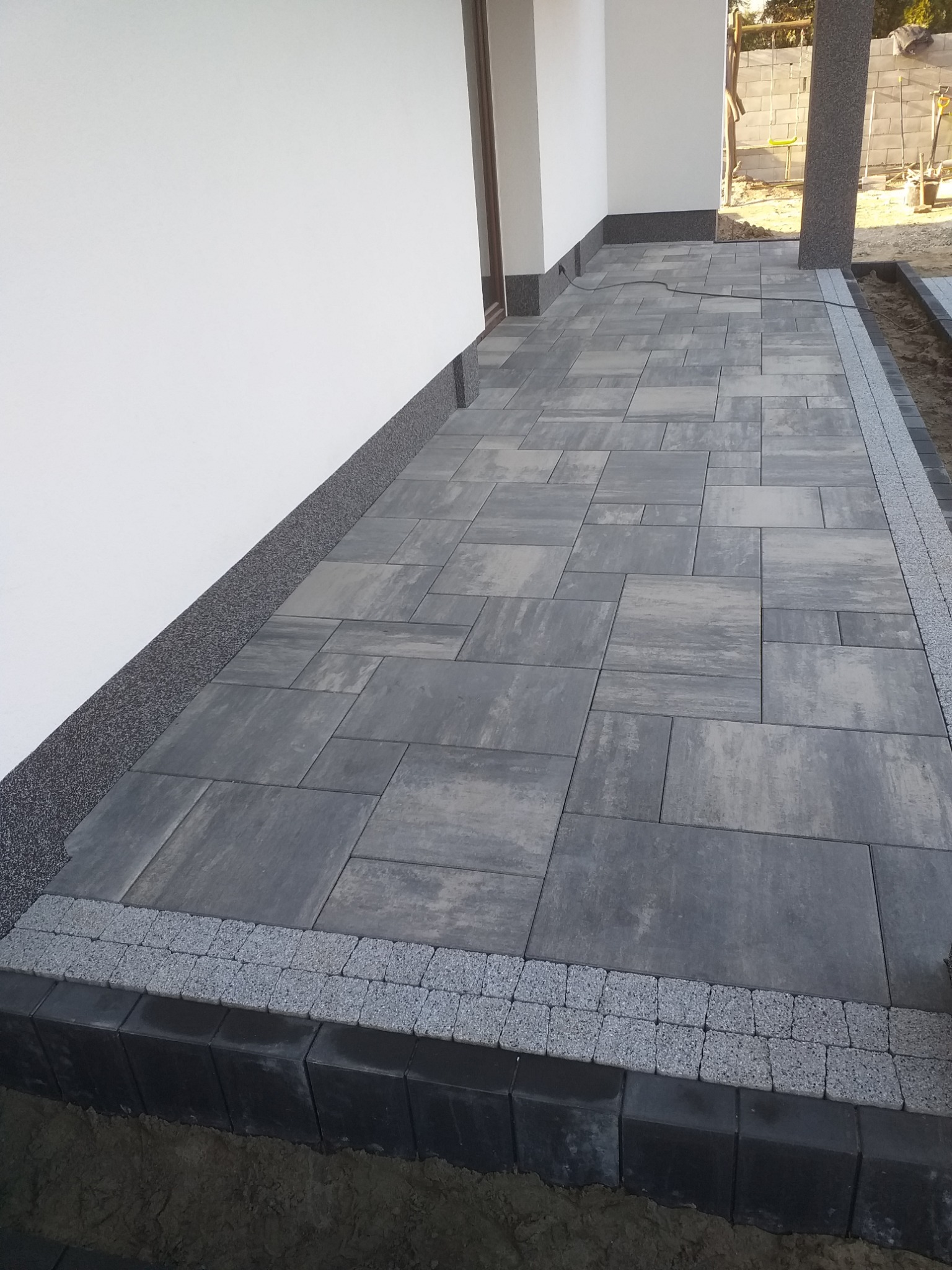 Ułożona szara kostka brukowa przed białym budynkiem, obrzeża z ciemnej kostki i jasnego grysu.