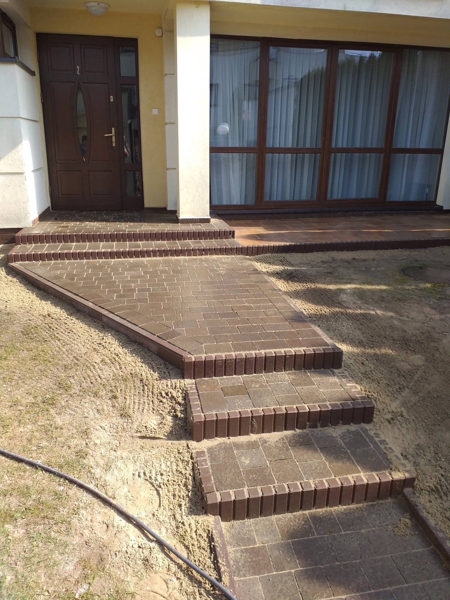 Schody wejściowe do domu wyłożone brązową kostką brukową, widoczne ślady piasku na trawniku.