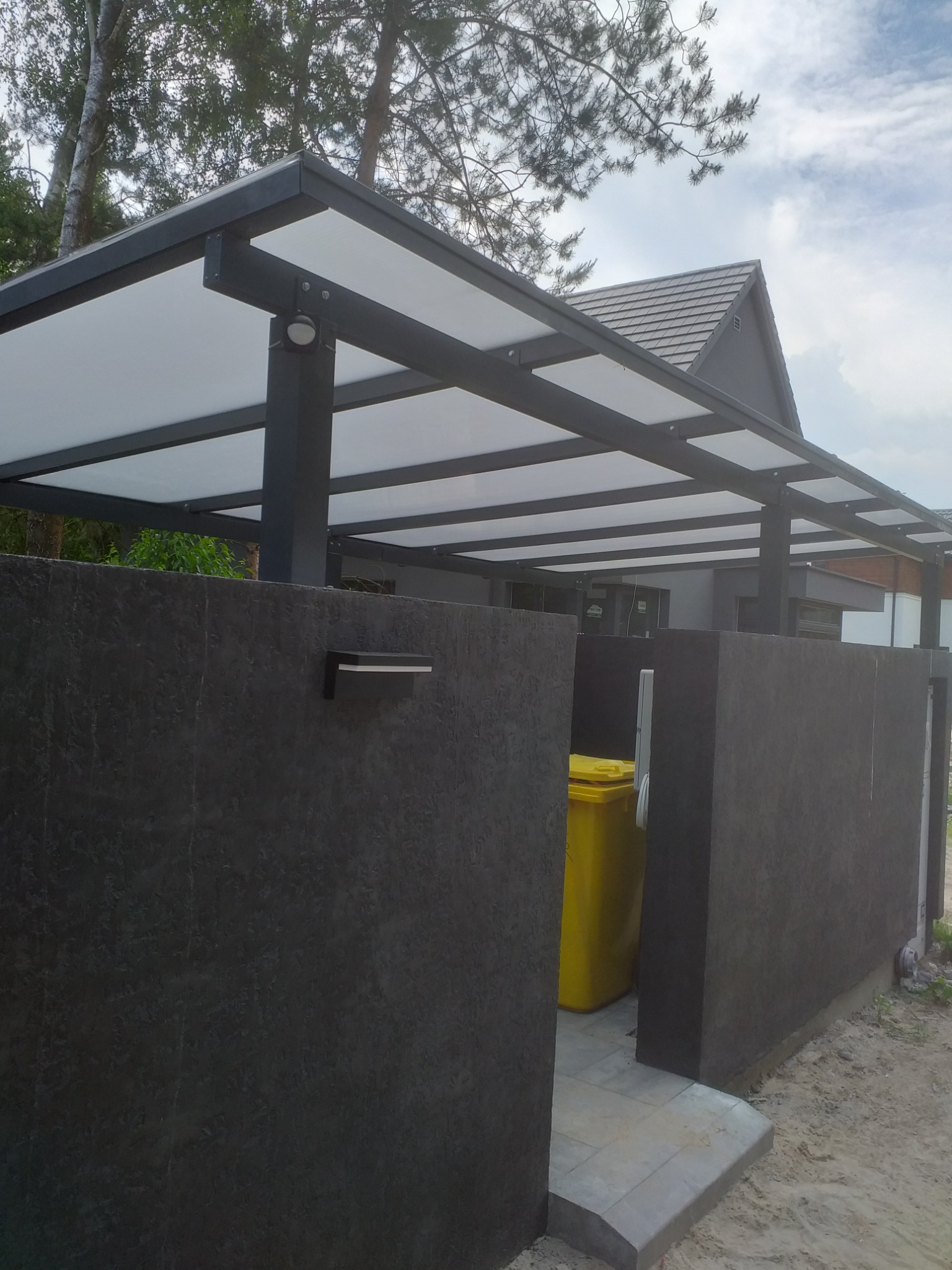 Nowoczesna, szara pergola z białym zadaszeniem nad wejściem do domu, otoczona ciemnoszarym murem z lampą i żółtym pojemnikiem na śmieci.