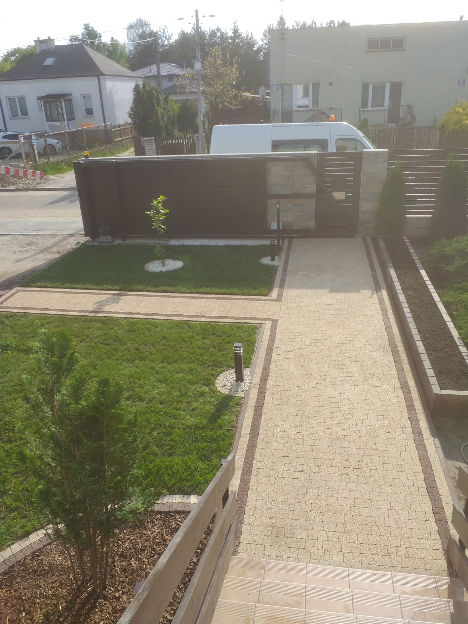 Widok z góry na nowo wybrukowaną ścieżkę z kostki w odcieniach beżu i brązu, prowadzącą do nowoczesnego, ciemnego ogrodzenia z furtką, z trawnikiem i młodymi drzewkami po bokach, w tle budynki...