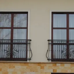 Dwa brązowe okna z ozdobnymi, kutymi balustradami, nad kamienną elewacją budynku w jasnych barwach. W szybach okien odbija się drzewo.