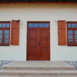 Elewacja domu z brązowymi oknami i okiennicami oraz dwuskrzydłowymi drzwiami wejściowymi, widoczne trzy stopnie schodów z kamienną podmurówką.