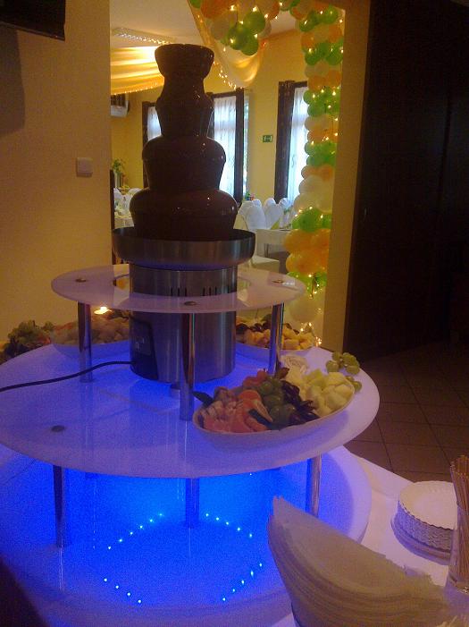 Fontanna czekoladowa na podświetlanym niebieskim stole z owocami, w tle dekoracja balonowa i nakryte stoły.