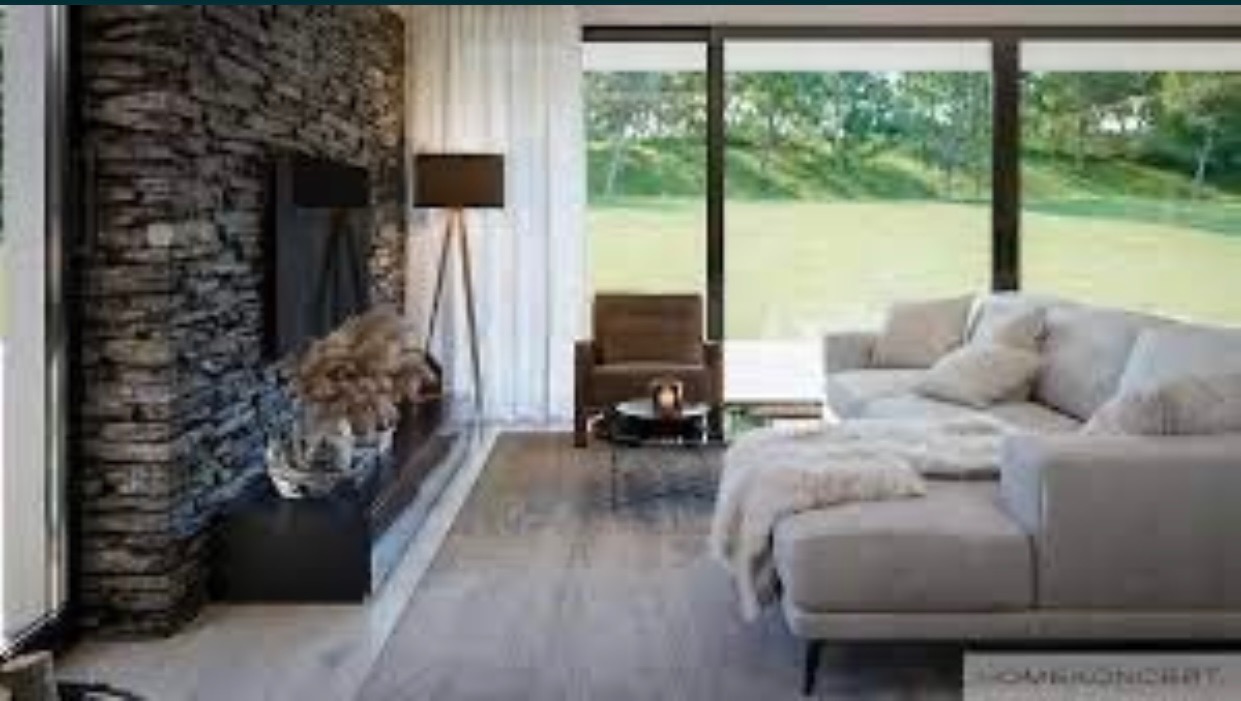 Nowoczesny salon z kamienną ścianą, szarą sofą narożną i dużym oknem z widokiem na zieleń. Na podłodze widoczna szara podłoga imitująca drewno. Czarna lampa stojąca i fotel dopełniają aranżację.