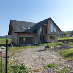 Dwukondygnacyjny dom w stanie surowym otwartym z czerwoną cegłą, czarnym dachem z oknami dachowymi, trójkątnym oknem na szczycie i taczką na żwirowym podjeździe w słoneczny dzień.