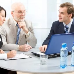 JADOTA KREDYTU - Spotkanie biznesowe: kobieta sporządza notatki, mężczyzna z brodą zamyślony, a drugi mężczyzna prezentuje coś na laptopie, na stole woda i szklanka.