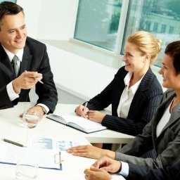 JADOTA KREDYTU - Grupa pięciu osób w garniturach siedzi przy białym stole podczas spotkania biznesowego, analizując wykresy i notując w notatniku.