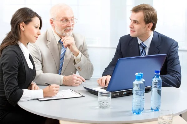 Spotkanie biznesowe: kobieta sporządza notatki, mężczyzna z brodą zamyślony, a drugi mężczyzna prezentuje coś na laptopie, na stole woda i szklanka.