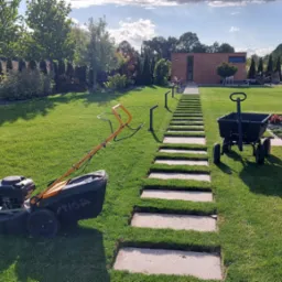 Żółto-czarna kosiarka Stiga na trawniku obok ścieżki z płyt betonowych prowadzącej do nowoczesnego domu, w tle zadbany ogród z drzewami i krzewami.