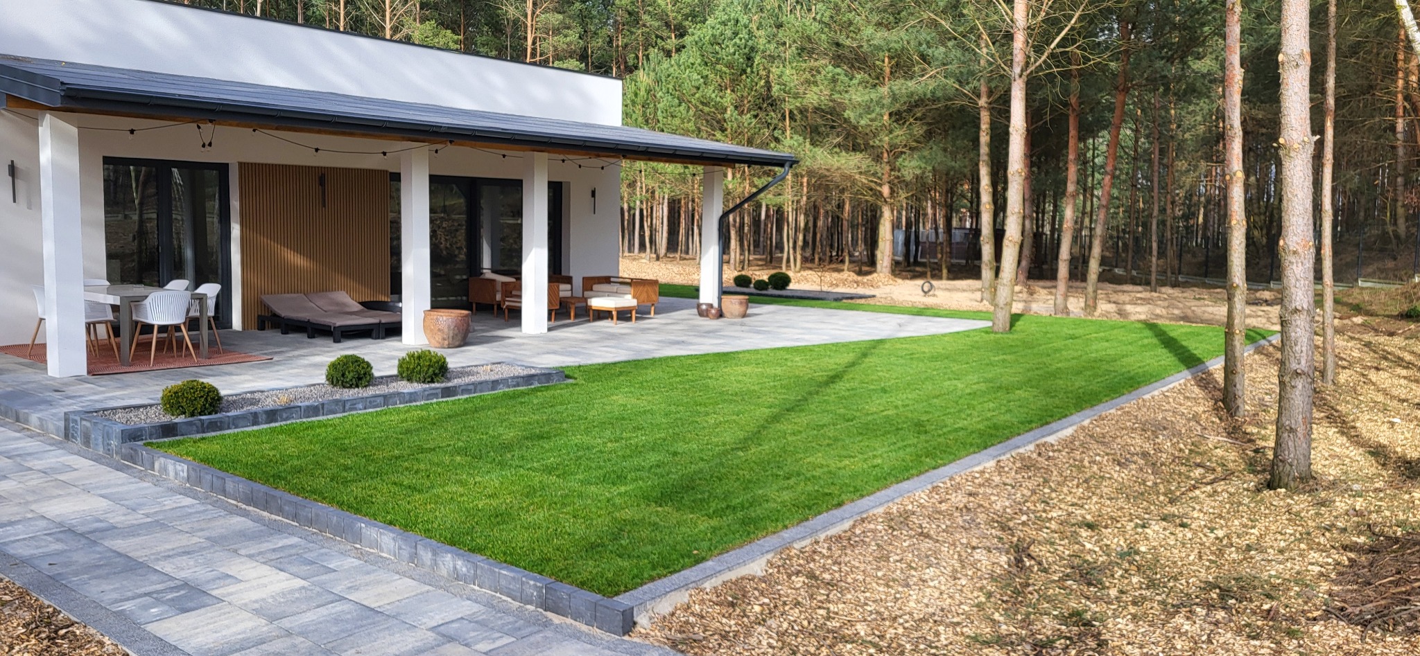 Nowoczesny dom z zadbanym trawnikiem, kostką brukową i elementami małej architektury ogrodowej w otoczeniu drzew.