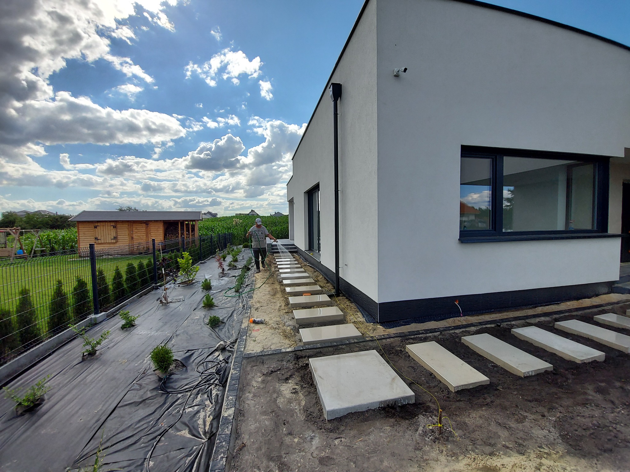 Nowoczesny dom z ogrodem w trakcie zakładania: widoczne sadzonki na agrowłókninie, ścieżka z płyt betonowych oraz osoba podlewająca rośliny w pobliżu budynku.