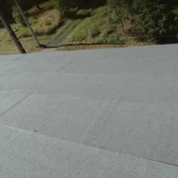 Szary, płaski dach pokryty papą termozgrzewalną, widok z góry na krawędź dachu i otoczenie z zielenią i słupem energetycznym.