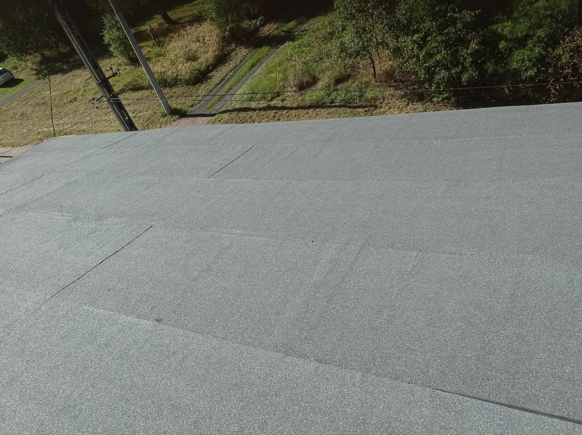 Szary, płaski dach pokryty papą termozgrzewalną, widok z góry na krawędź dachu i otoczenie z zielenią i słupem energetycznym.