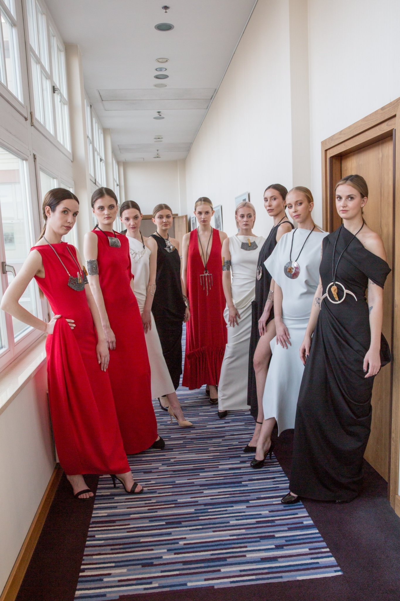 Grupa ośmiu modelek prezentuje kolekcję sukienek w odcieniach czerwieni, bieli i czerni, każda z unikalną biżuterią, w jasnym korytarzu z oknami i wzorzystą wykładziną.