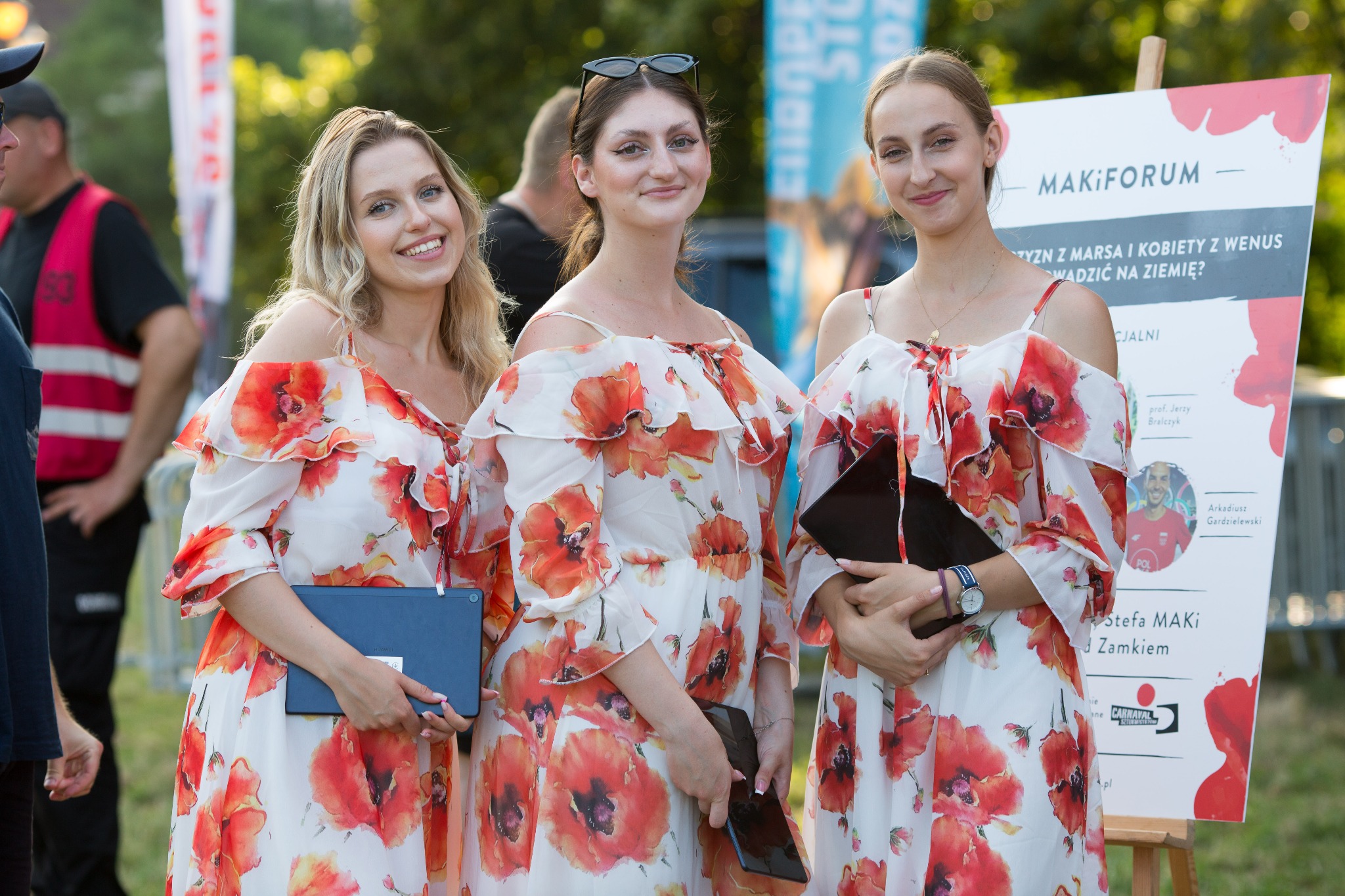 Trzy uśmiechnięte hostessy w sukienkach w makowe wzory, trzymające tablety, stojące obok tablicy informacyjnej Maki Forum podczas eventu plenerowego.