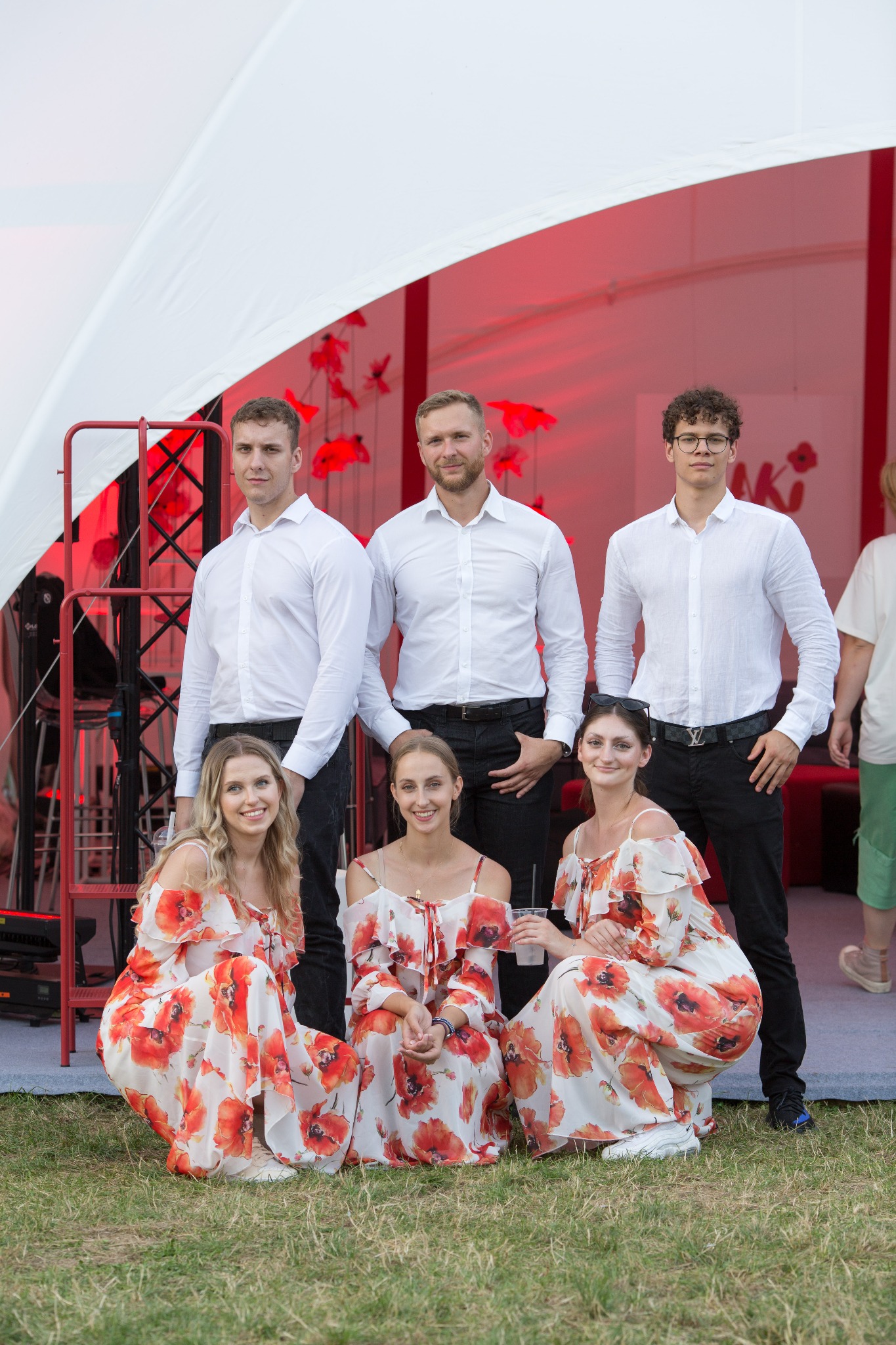 Grupa trzech mężczyzn w białych koszulach i trzech kobiet w sukienkach w makowy wzór pozuje na tle białego namiotu z czerwonymi dekoracjami.