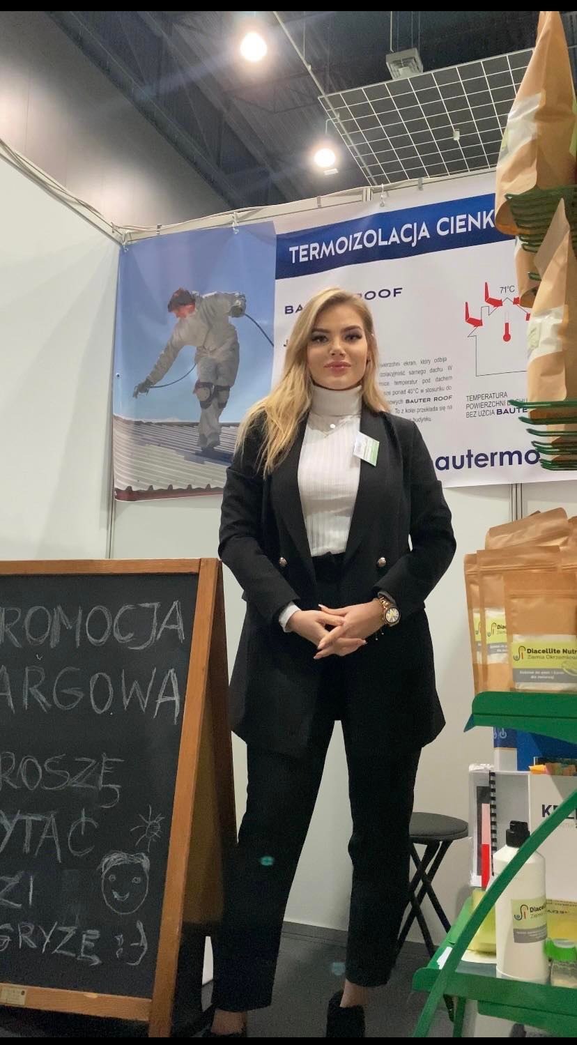 Kobieta w czarnym garniturze prezentuje się na tle stoiska targowego z materiałami do termoizolacji cienkowarstwowej, obok tablica z napisem 'Promocja Targowa' i rysunkiem uśmiechniętej buzi.