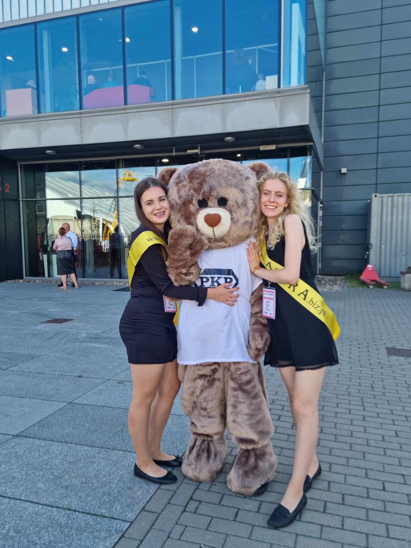 Dwie uśmiechnięte kobiety w czarnych sukienkach i szarfach z napisem, przytulają pluszowego misia w białej koszulce z logo PKP na tle nowoczesnego budynku z dużymi oknami.