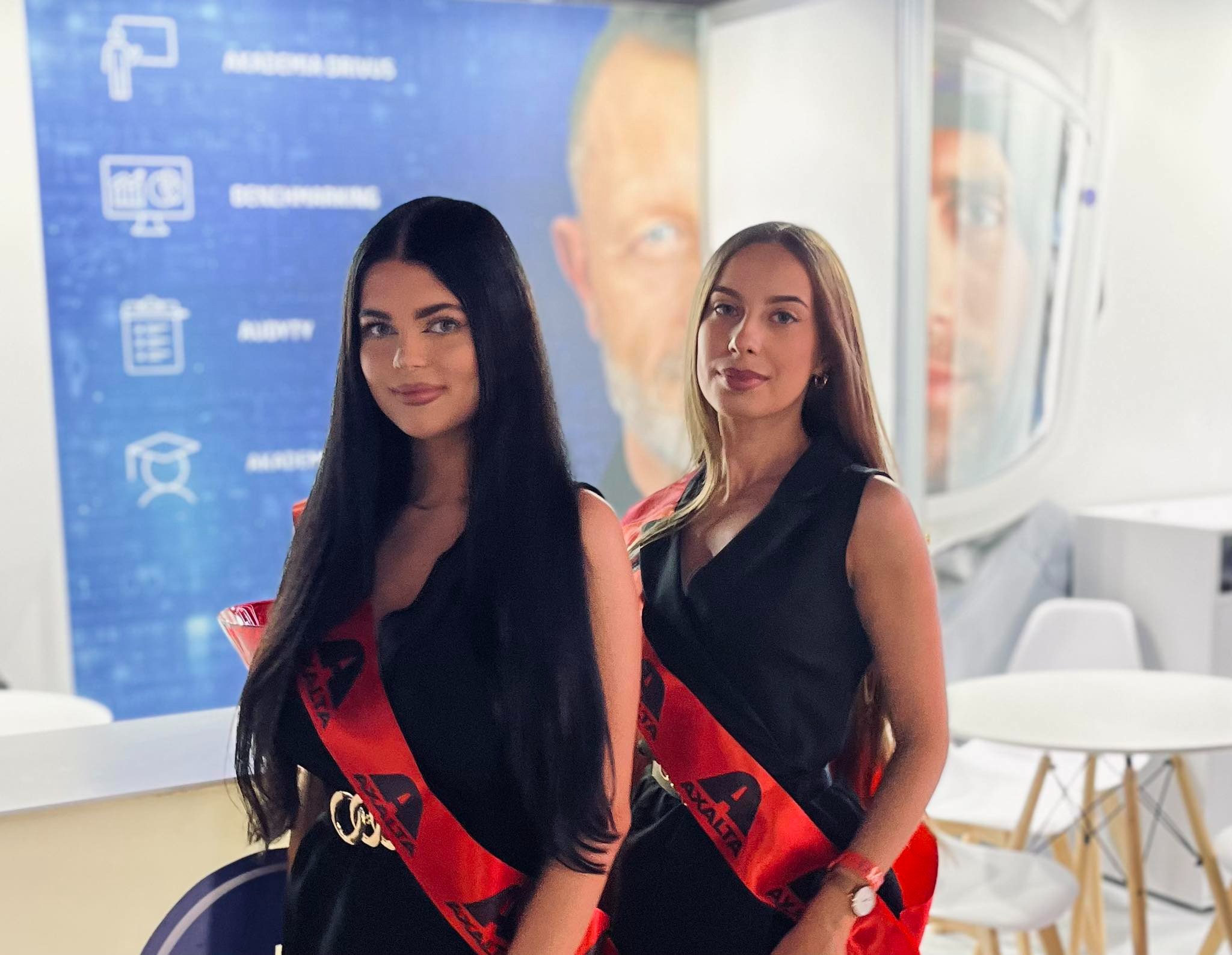 Dwie uśmiechnięte hostessy w czarnych sukienkach i czerwonych szarfach z logo firmy Axalta, stojące na tle stoiska targowego z grafikami i portretem mężczyzny.