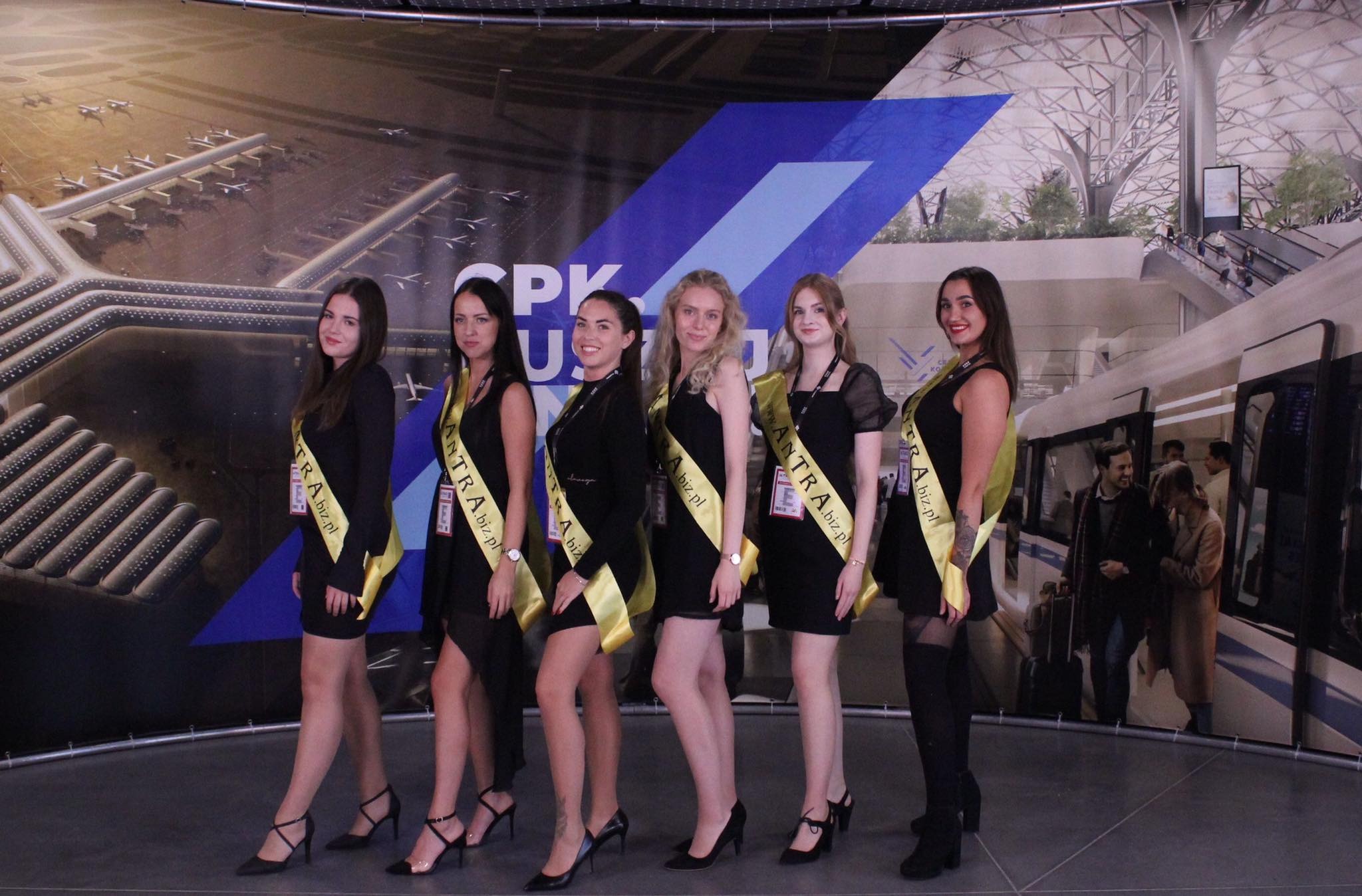Sześć młodych kobiet w czarnych sukienkach i szarfach z napisem ANTRA.biz.pl pozuje na tle grafiki przedstawiającej lotnisko i pociąg.
