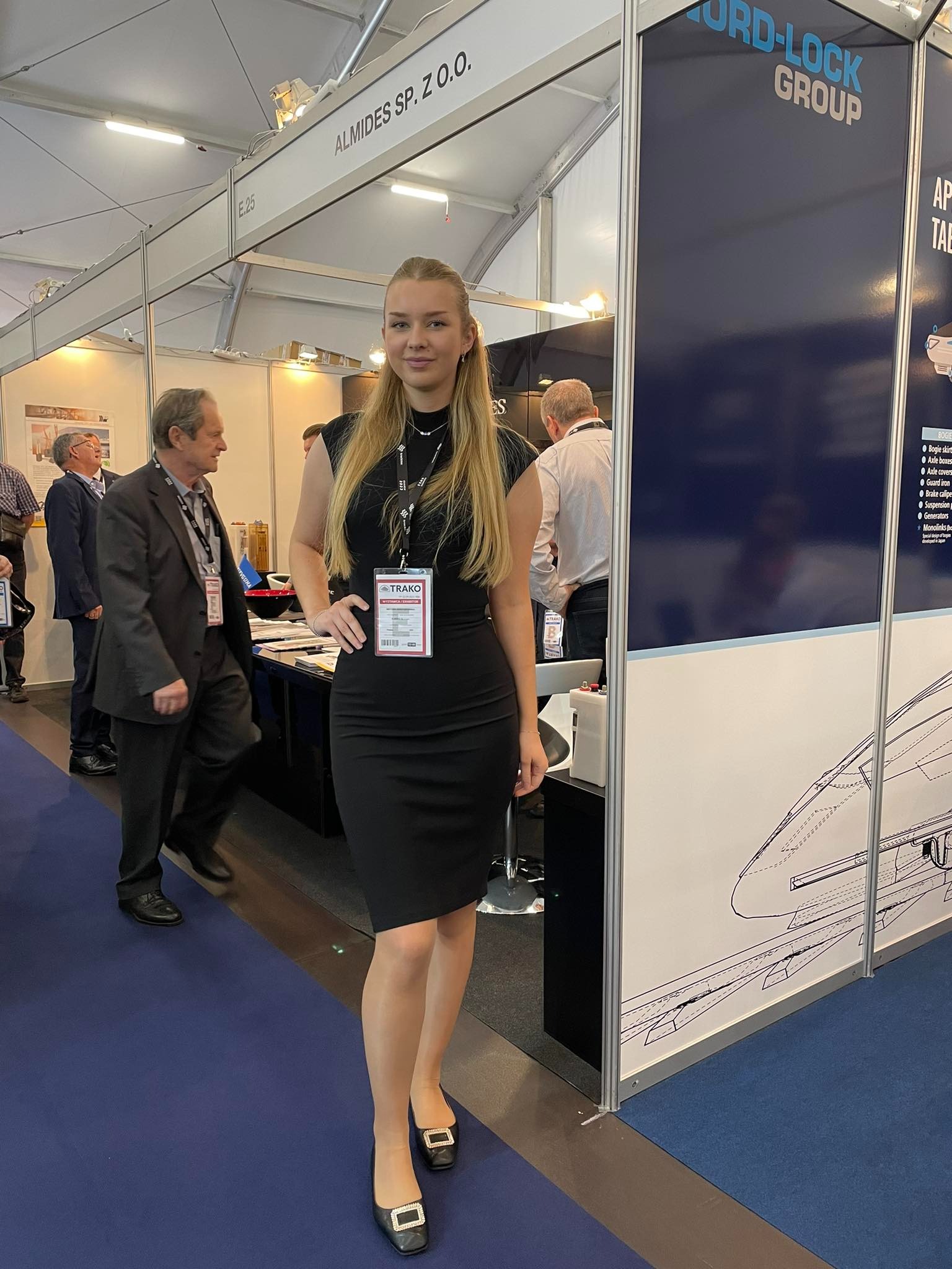 Kobieta w czarnej sukience pozuje na tle stoiska firmy AlMIDES SP. Z O.O. na targach TRAKO, z widocznym logo NORD-LOCK GROUP i schematem pociągu.