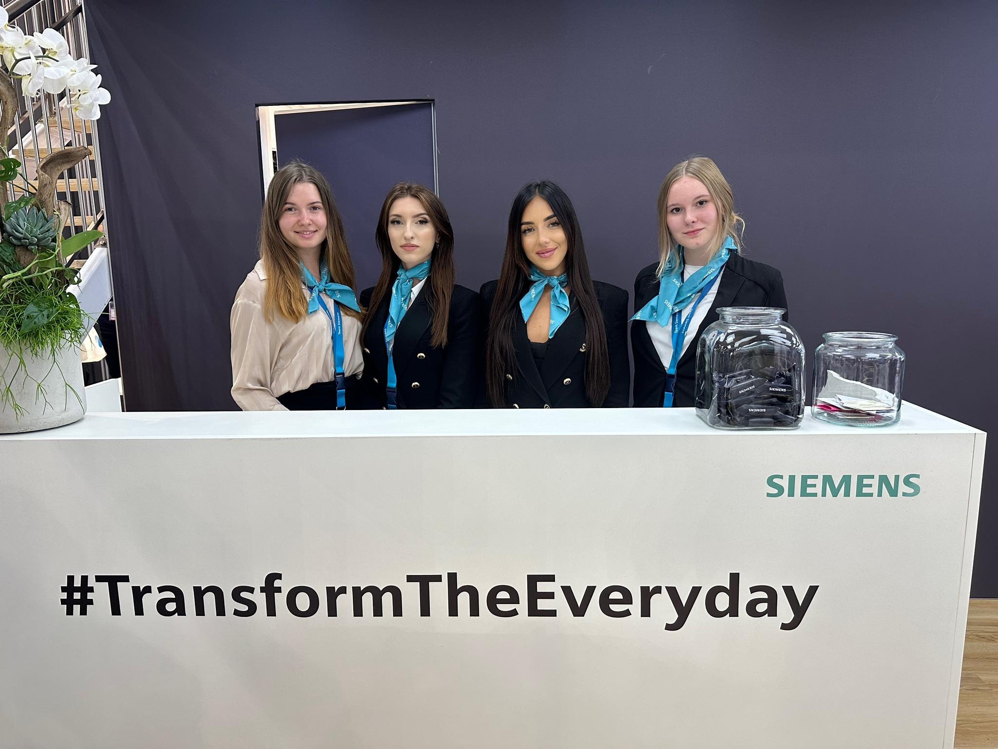 Cztery kobiety ubrane w stroje biznesowe z turkusowymi apaszkami stoją za białym stoiskiem z napisem #TransformTheEveryday i logo Siemens, obok dekoracji z białych kwiatów i sukulentów, na tle...