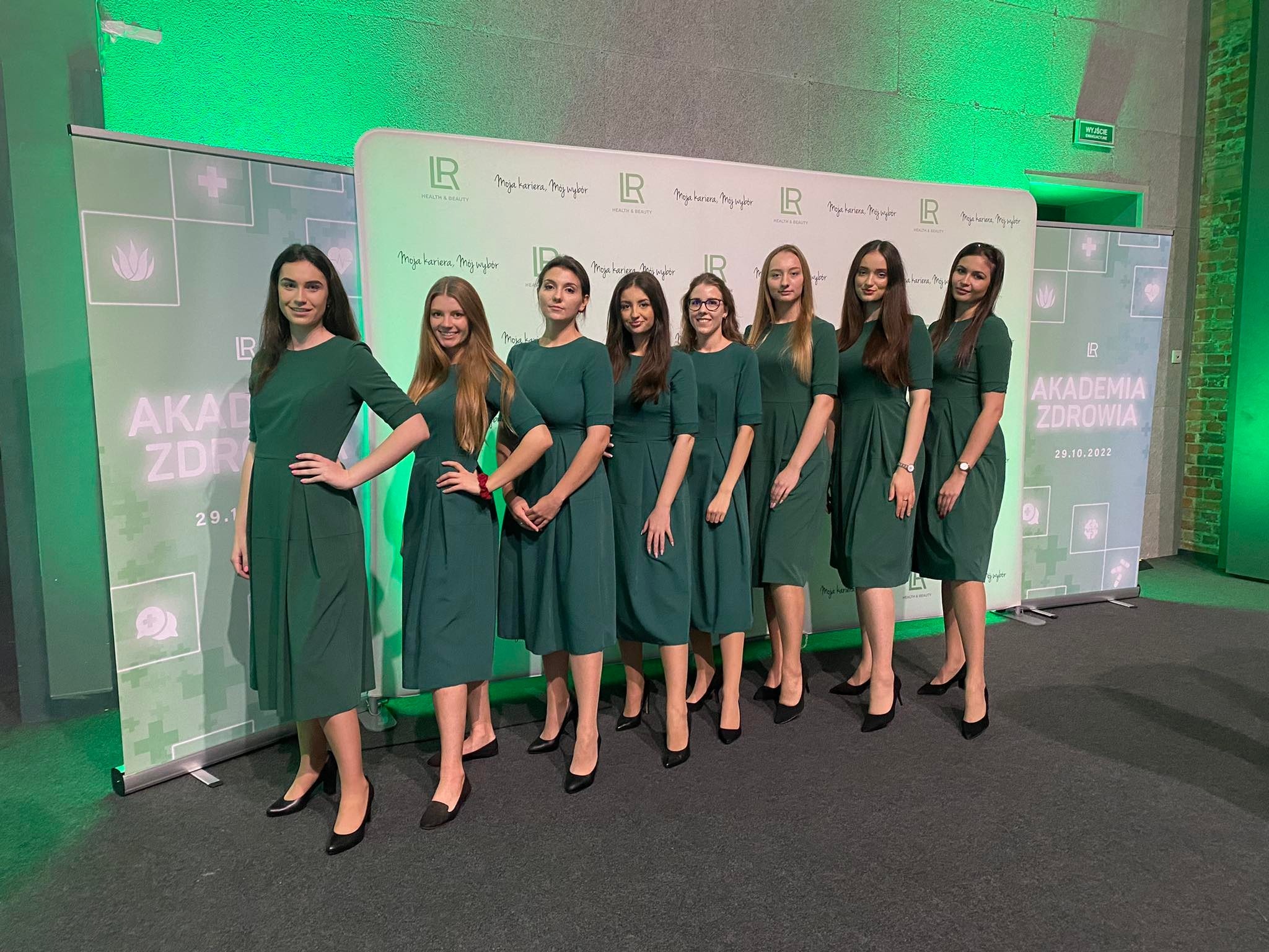 Grupa ośmiu kobiet w zielonych sukienkach pozuje na tle roll-upu z logiem i napisem 'Akademia Zdrowia', czarne szpilki, zielone oświetlenie.