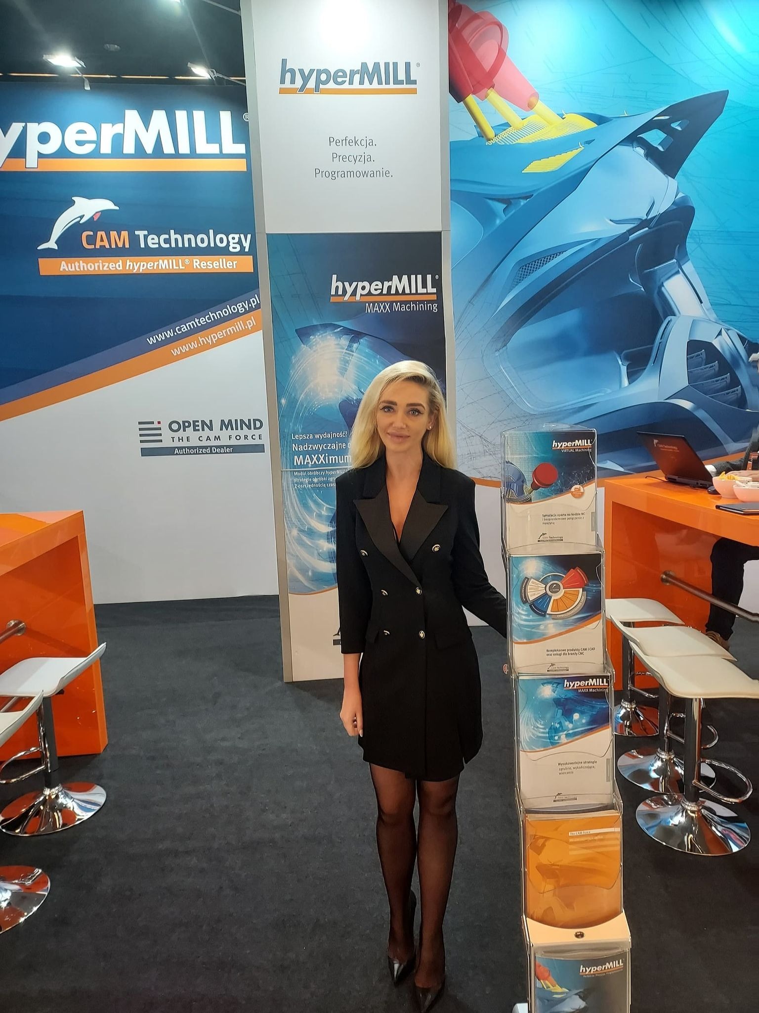 Kobieta w czarnej sukience na stoisku targowym HyperMill z materiałami reklamowymi CAM Technology i Open Mind, prezentującymi oprogramowanie do obróbki CNC.