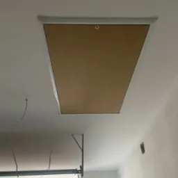 Zamknięty wyłaz strychowy z brązową klapą i białym obramowaniem w białym suficie, widoczne fragmenty drabiny i instalacji elektrycznej.