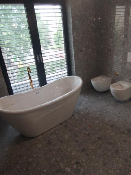 Nowoczesna łazienka z wolnostojącą białą wanną przy oknie z żaluzjami i podwieszanymi toaletami na tle ściany z szarego kamienia.