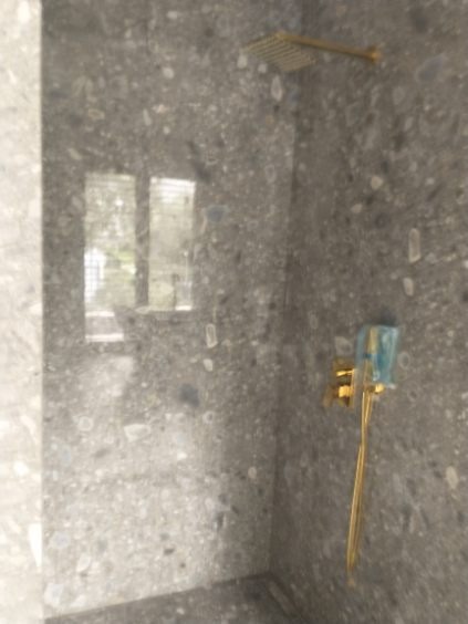 Wykończona łazienka z szarą, wzorzystą okładziną ścian, złotą deszczownicą i baterią, widoczna szyba.