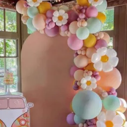 Pastelowa girlanda z balonów i kwiatów, tworząca łuk na tle jasnej ściany, z kartonową dekoracją w kształcie busa w stylu retro z lewej strony, oraz tort w oknie w tle.