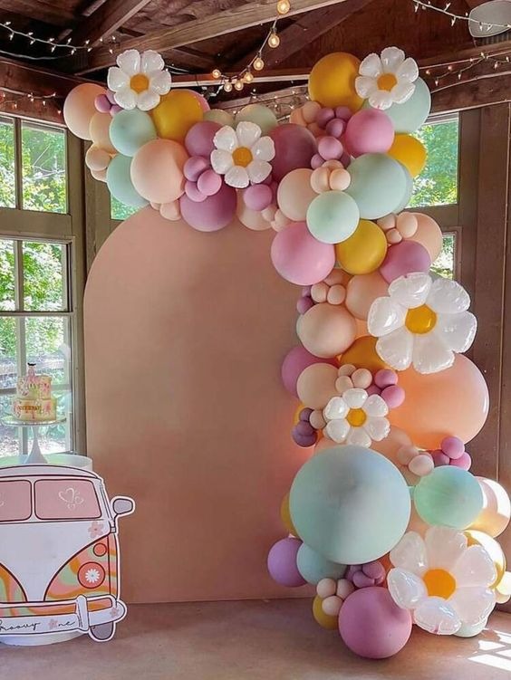 Pastelowa girlanda z balonów i kwiatów, tworząca łuk na tle jasnej ściany, z kartonową dekoracją w kształcie busa w stylu retro z lewej strony, oraz tort w oknie w tle.