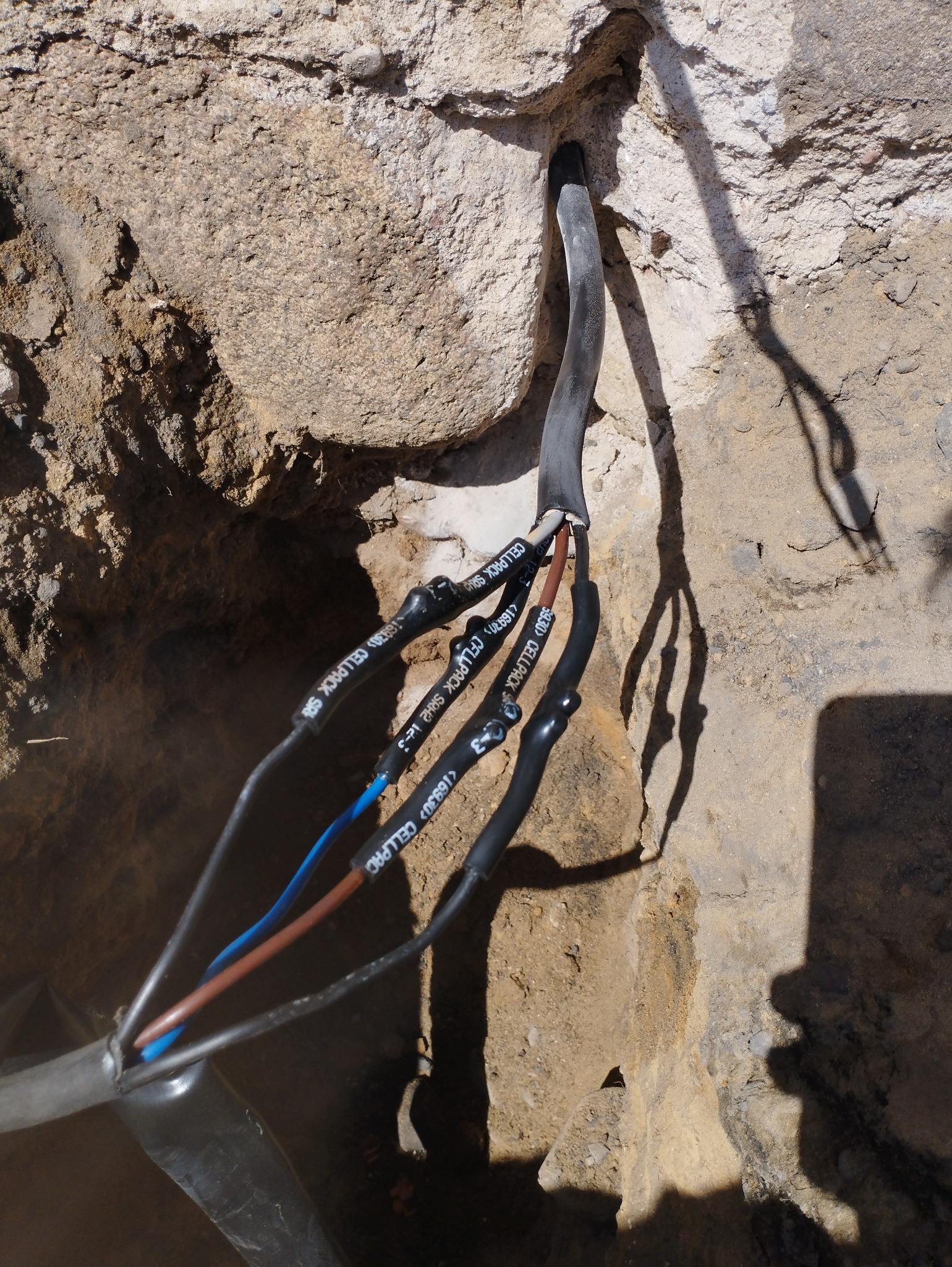 Kabel elektryczny z izolacją termokurczliwą Cellpack wychodzący z otworu w ścianie z betonu komórkowego, widoczne przewody w kolorach niebieskim, brązowym i czarnym.