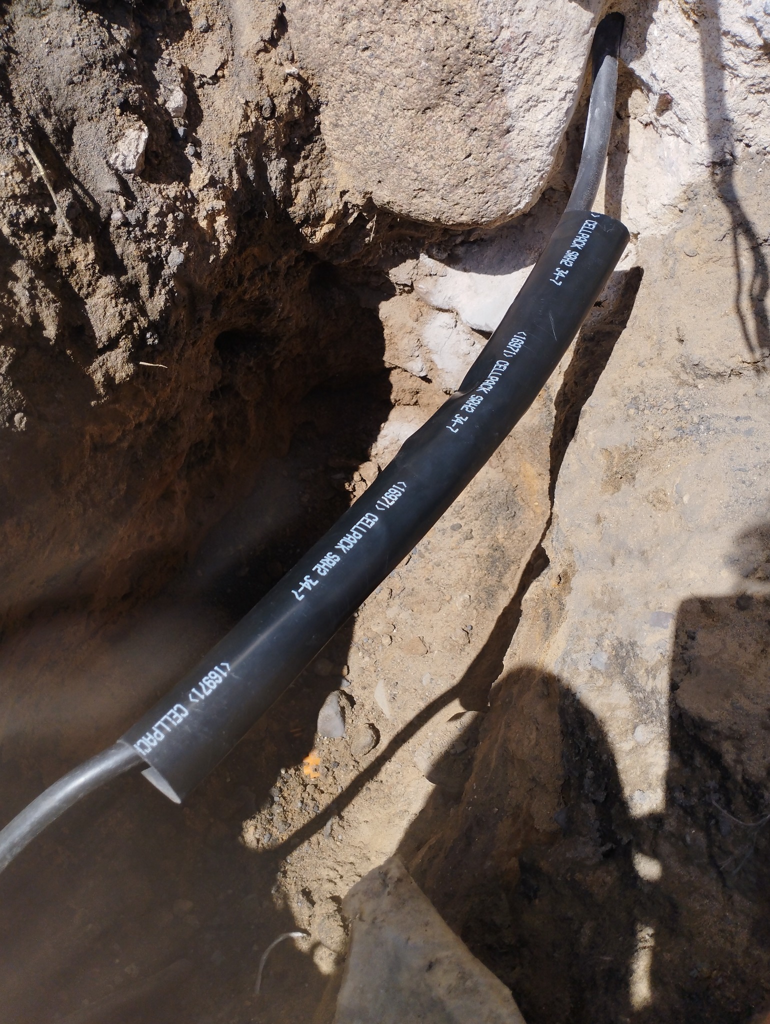 Kabel elektryczny w czarnej osłonie ochronnej CELLPACK SRH2 34-7 ułożony w wykopie ziemnym, widoczne kamienie i piasek.
