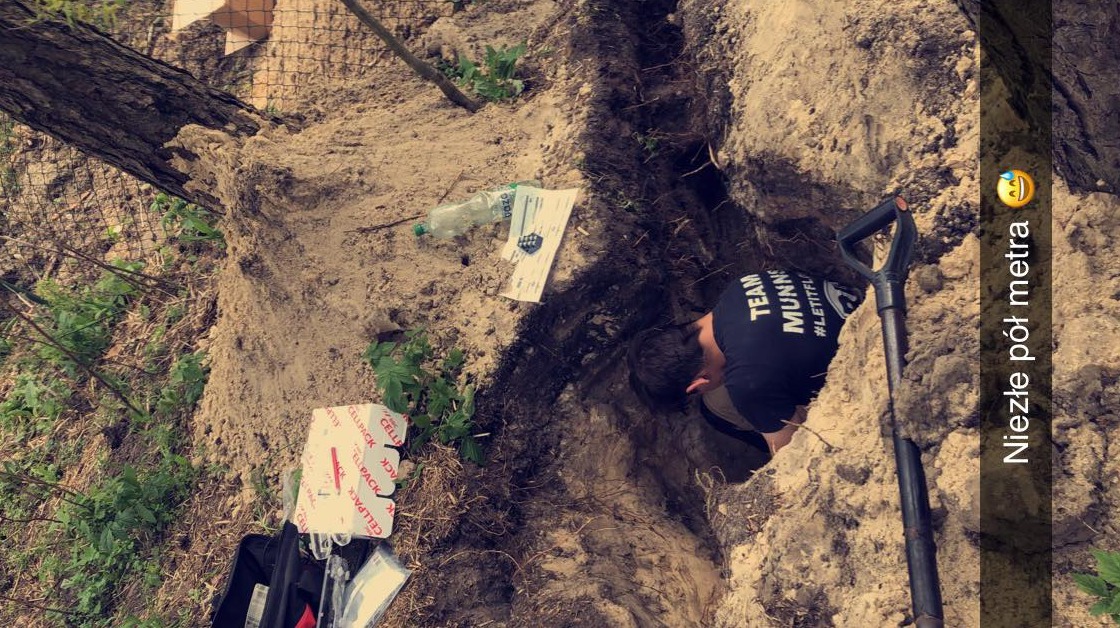 Mężczyzna w czarnej koszulce kopie wąski rów w ziemi, obok leżą śmieci i narzędzia, widoczna łopata wbita w ziemię.