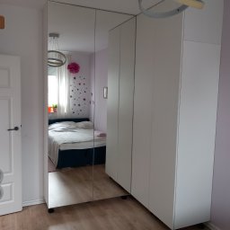 Kacper Siwek Studio Mebli - Biała szafa z lustrzanymi drzwiami w sypialni, odbijająca łóżko i okno, oświetlona nowoczesną lampą sufitową.