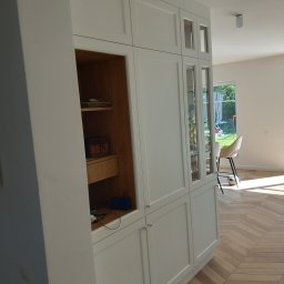 Kacper Siwek Studio Mebli - Biała zabudowa meblowa z przeszklonymi witrynami i otwartą półką z drewnianym wnętrzem, na podłodze parkiet w jodełkę, w tle widok na ogród przez okno.