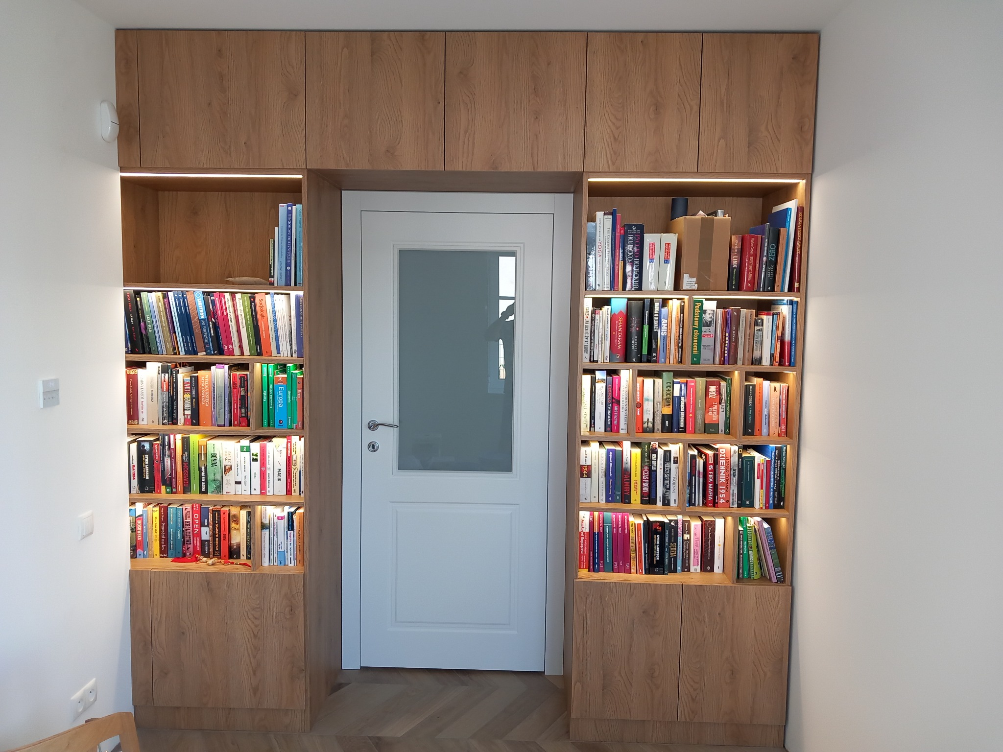 Zabudowa wnęki z półkami na książki, drewnopodobne fronty, oświetlenie LED, białe drzwi z szybą w centralnej części zabudowy, parkiet w jodełkę na podłodze.