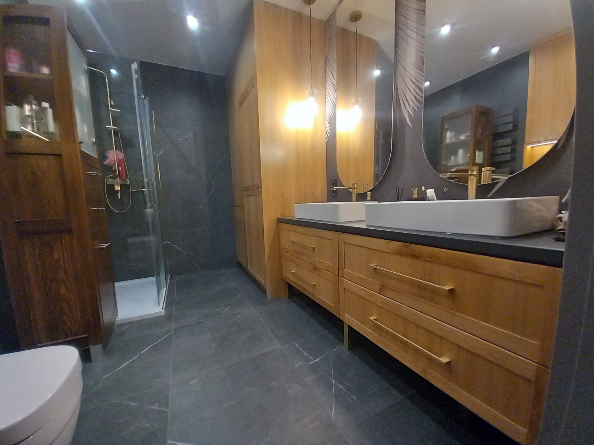 Nowoczesna łazienka z drewnianymi szafkami pod umywalki, dużym owalnym lustrem z dekoracyjnym nadrukiem, szklaną kabiną prysznicową i ciemnymi płytkami podłogowymi