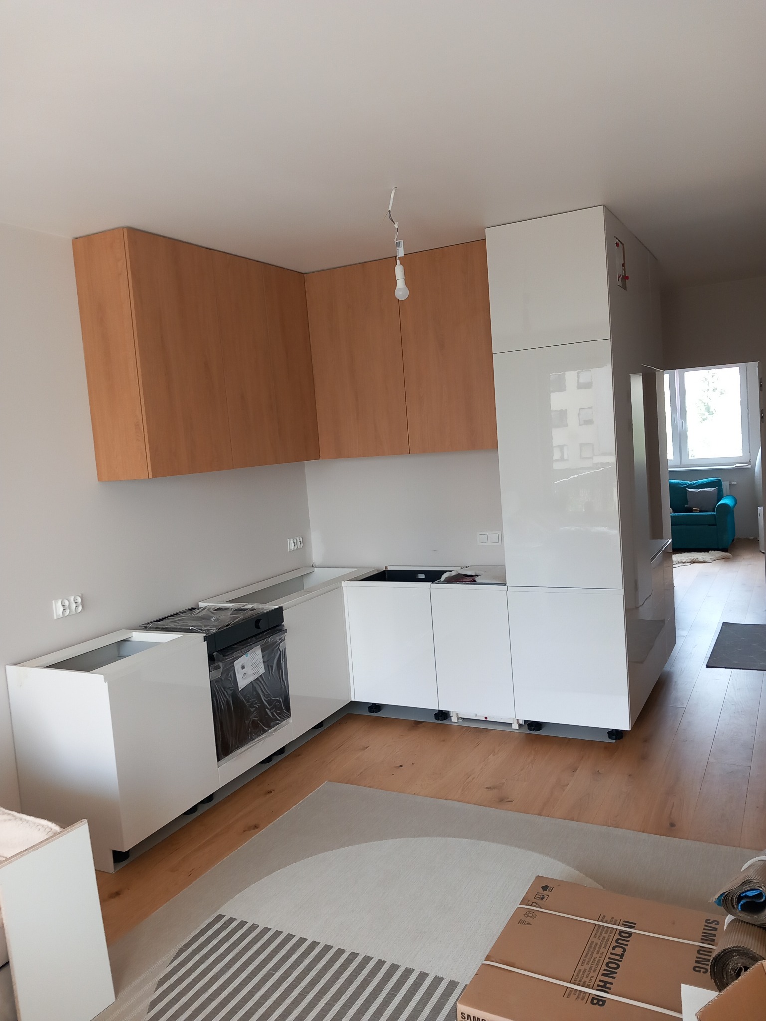 Montaż nowoczesnej kuchni: białe i drewniane szafki wiszące, płyta indukcyjna w folii ochronnej, widoczne elementy wykończeniowe na podłodze.