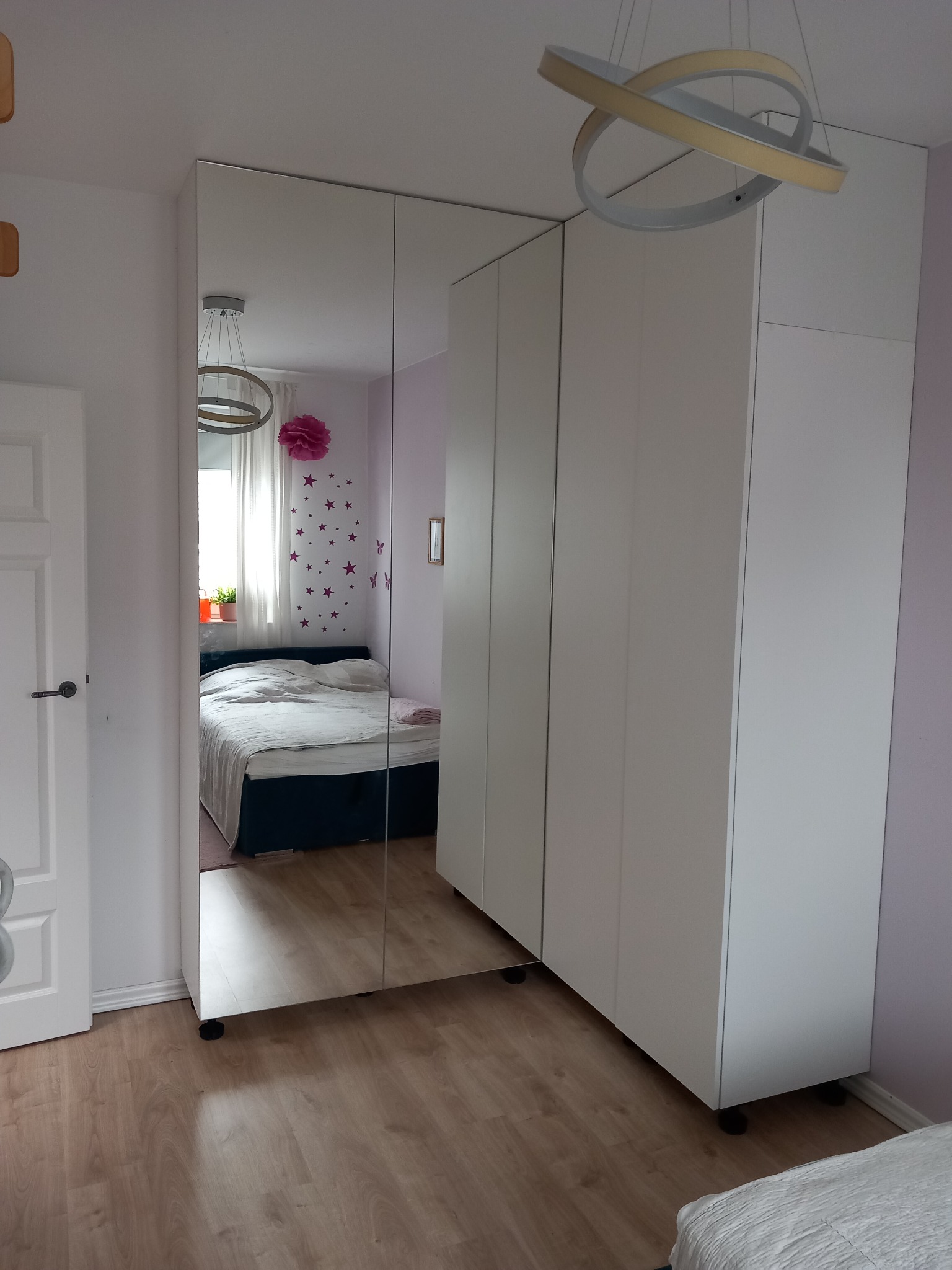 Biała szafa z lustrzanymi drzwiami w sypialni, odbijająca łóżko i okno, oświetlona nowoczesną lampą sufitową.